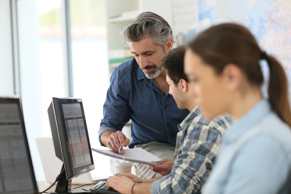 a female and male employee sit in front of computer monitors while an instructor leans in to explain a topic to them
